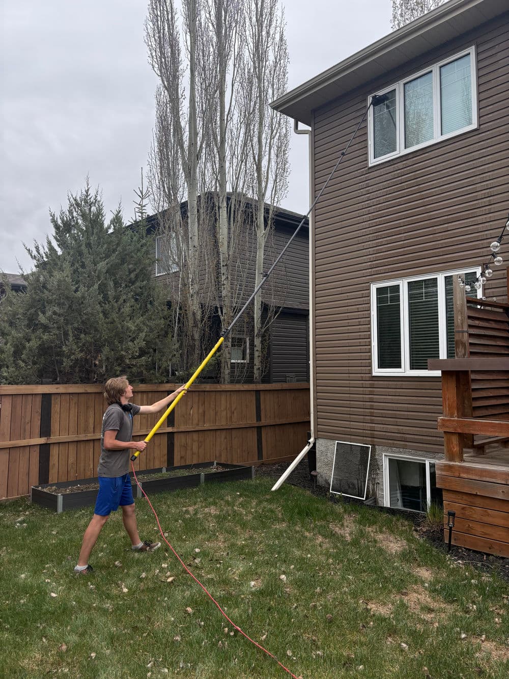 Person using a pole to clean windows on a two-story house with lush grass and trees.
