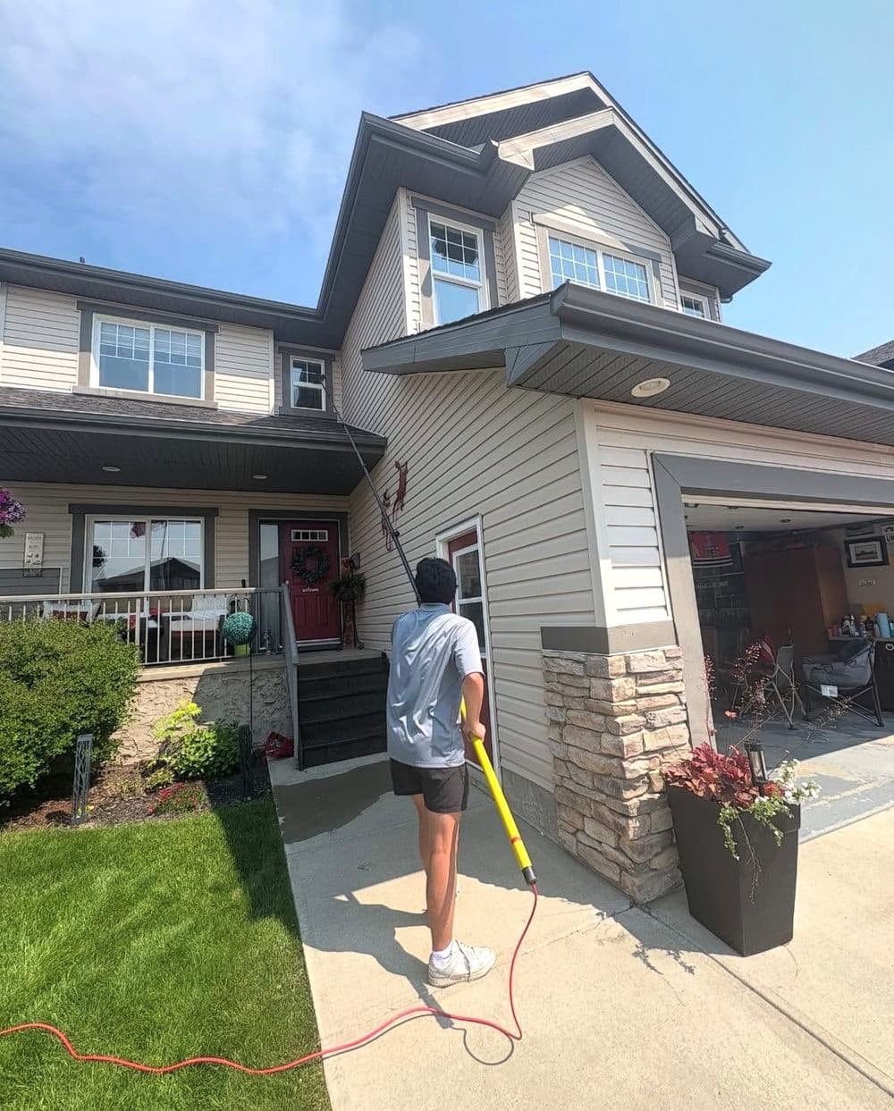 Person using a pressure washer to clean the exterior of a two-story house on a sunny day.