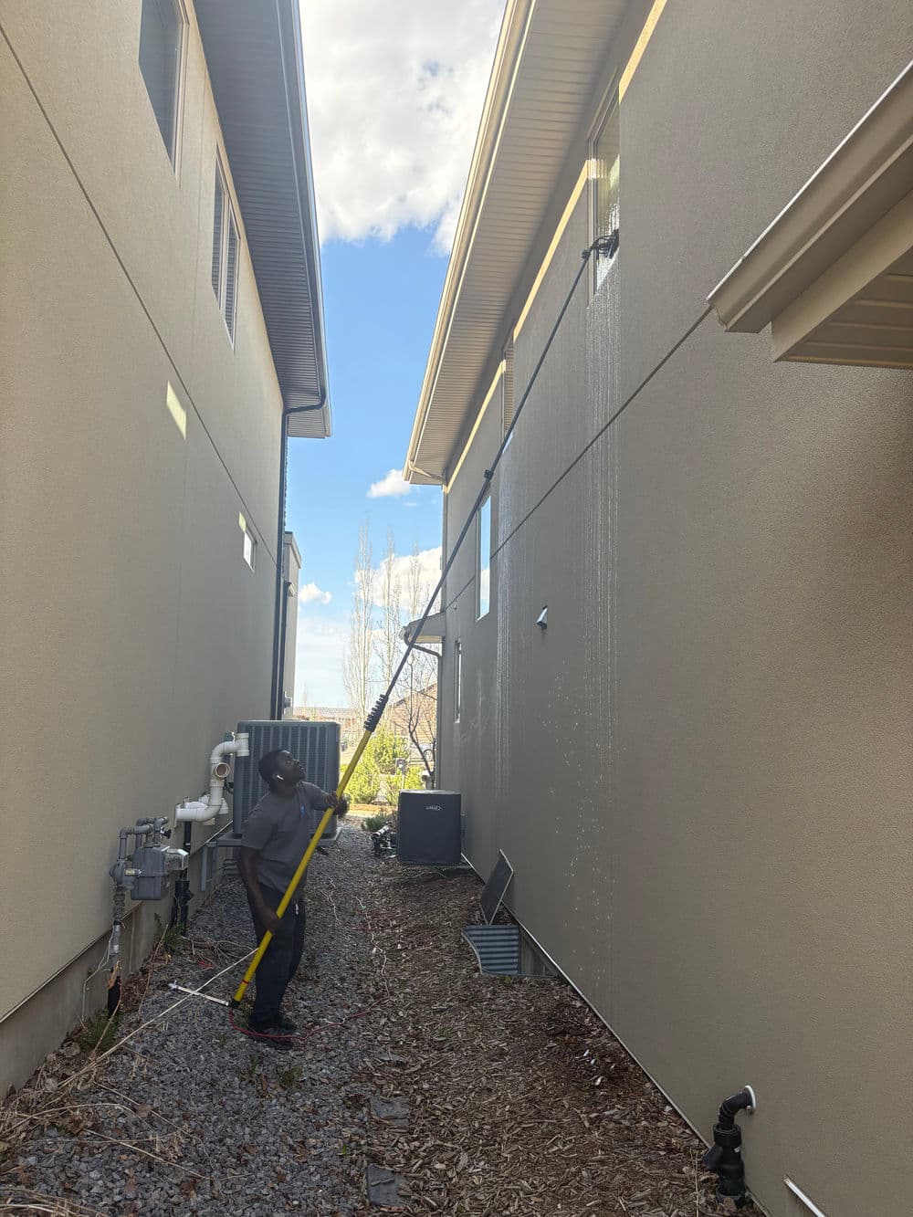 Person cleaning the side of a house with a long pole and hose under a blue sky.