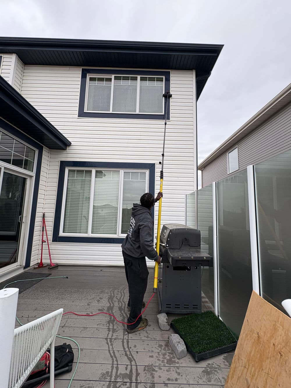 Person using a pole to clean the exterior of a house, with a grill and patio visible.