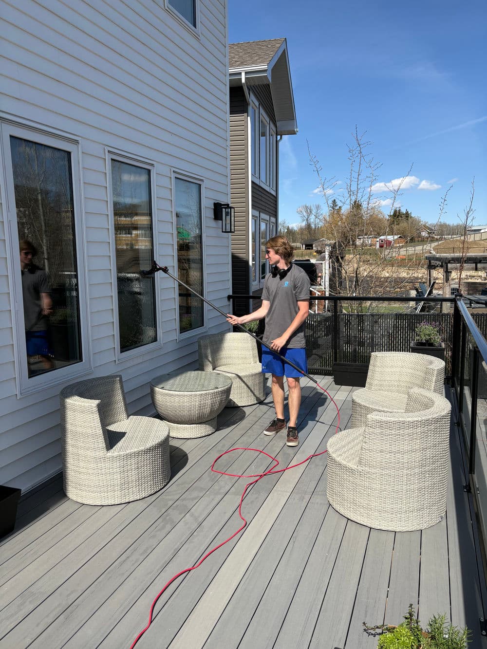 Person using a pressure washer to clean windows on a sunny outdoor patio with wicker furniture.