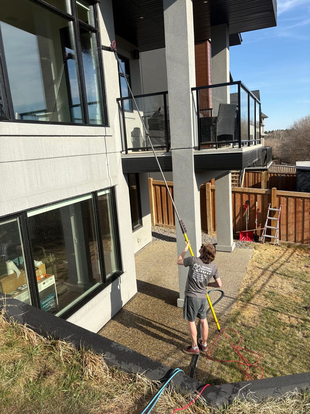 Man using a pole to clean high windows on a modern house, with outdoor furniture visible.