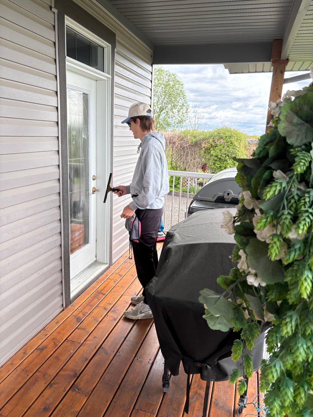 Person cleaning a glass door on a porch with a grill nearby. Bright exterior setting.
