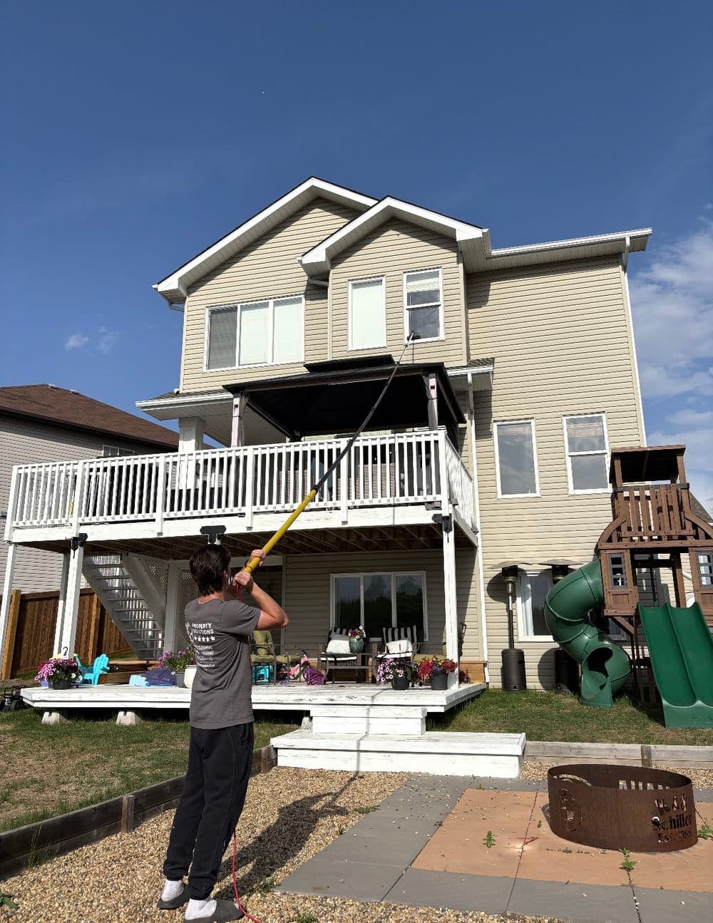 Person using a pole to wash windows on a multi-story house with outdoor play equipment.